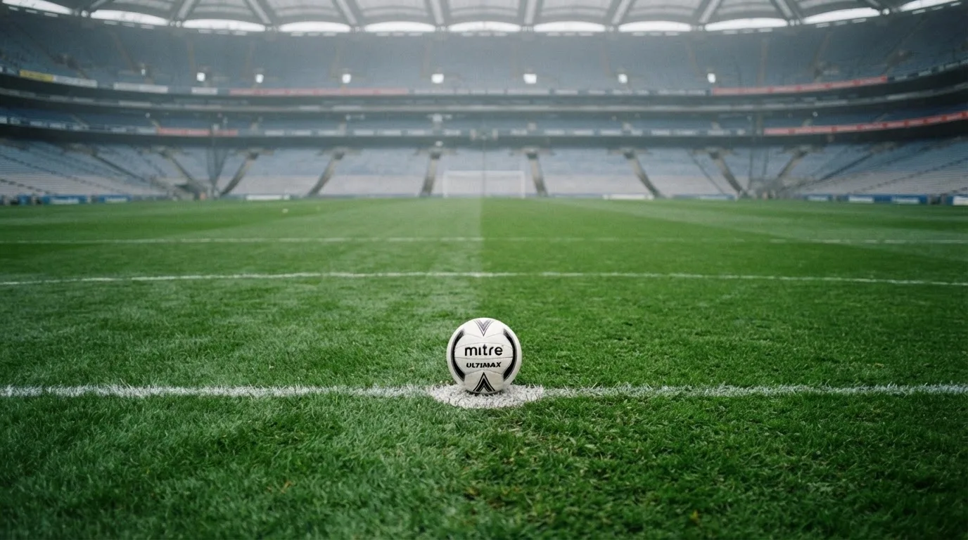 Ein Fußball liegt auf dem Elfmeterpunkt eines leeren Rasenplatzes, im Hintergrund eine Tribüne im Dunst.