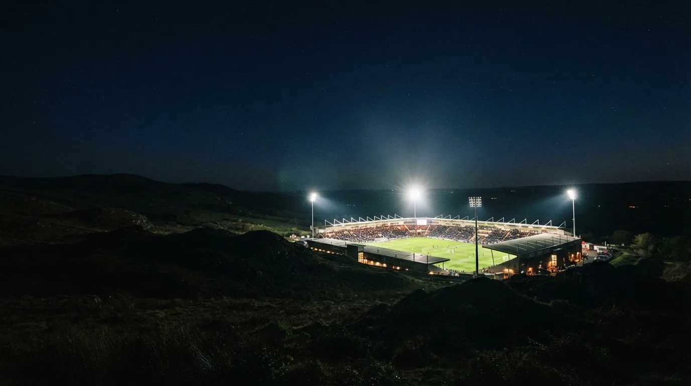 Ein Fußballstadion bei Nacht, aus der Ferne gesehen, Flutlicht über dem hell erleuchteten Rasen, davor dunkle Hügel.