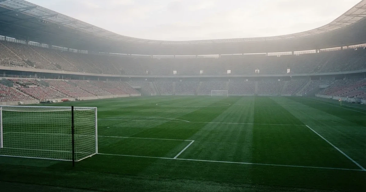 Ein Fußballplatz mit gepflegtem Rasen und einem Tor, über dem Spielfeld liegt Morgennebel, im Hintergrund eine leere Tribüne.
