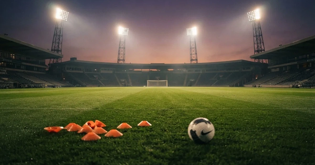 Eine Flutlichtanlage ragt über einen Trainingsplatz, auf dem Rasen liegen verstreut Hütchen und ein Ball, am Himmel die Abenddämmerung.