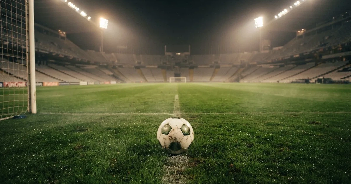 Ein Fußball liegt auf dem Rasen eines leeren Stadions bei Flutlicht, im Hintergrund unscharfe Tribünen ohne erkennbare Beschriftung.
