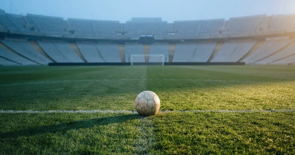 Ein Fußball liegt auf dem Mittelkreis eines Stadionrasens am frühen Morgen, im Hintergrund leere Ränge ohne erkennbare Beschriftung.