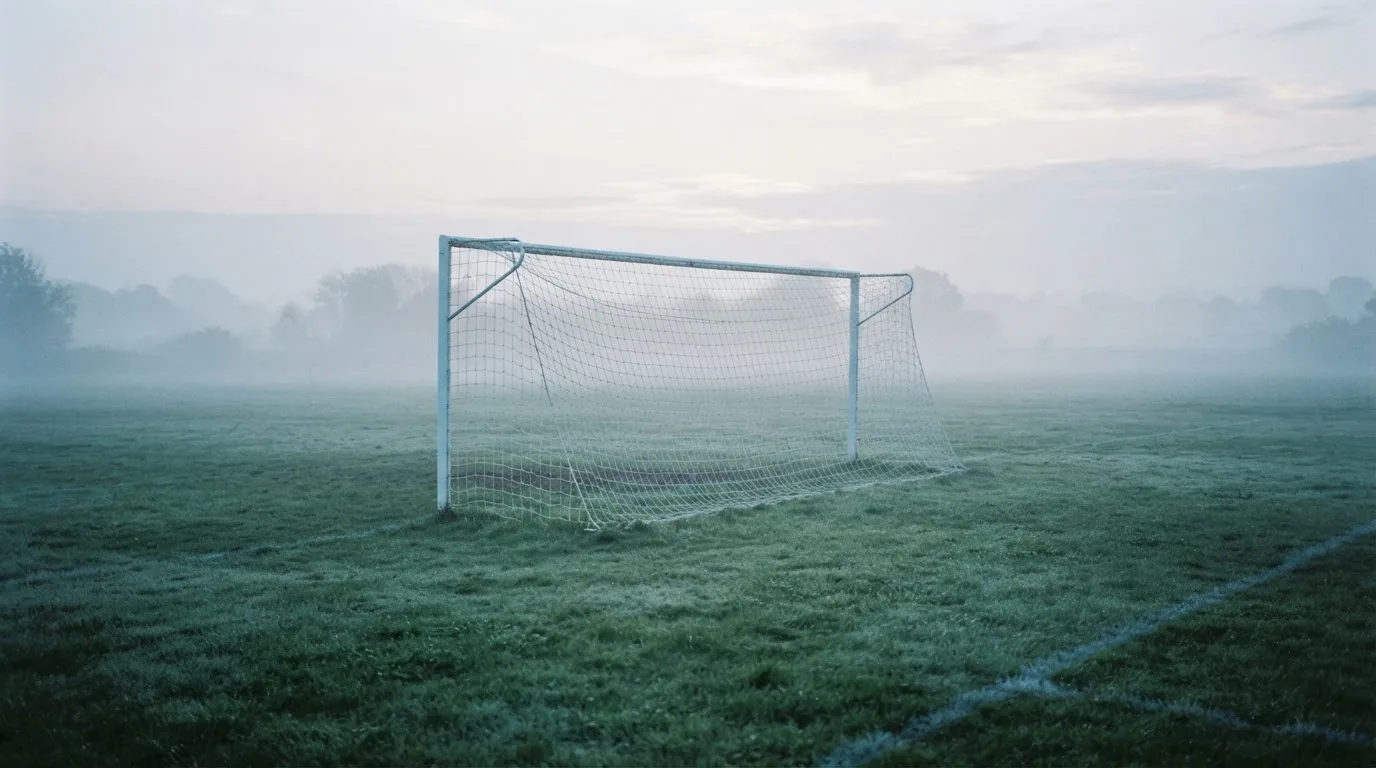 Ein Fußballtor mit Netz im Morgennebel auf einem leeren Platz, die Linien nur schwach zu erkennen.