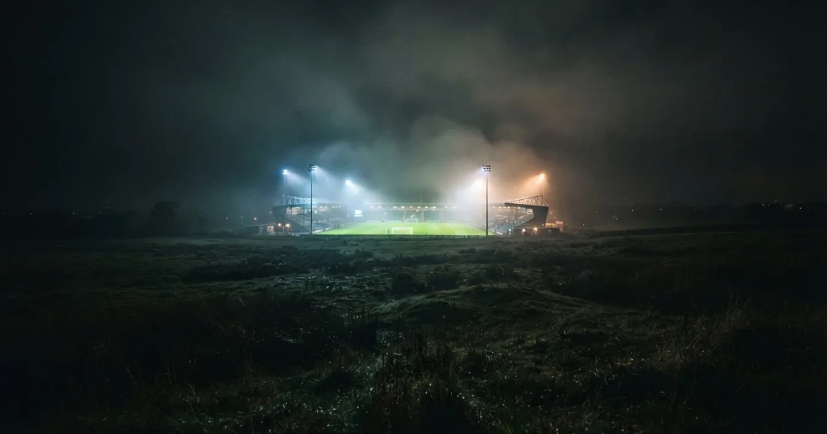 Ein Stadion unter Flutlicht, aus großer Entfernung über eine dunkle Wiese fotografiert, am Himmel zieht Nebel auf.