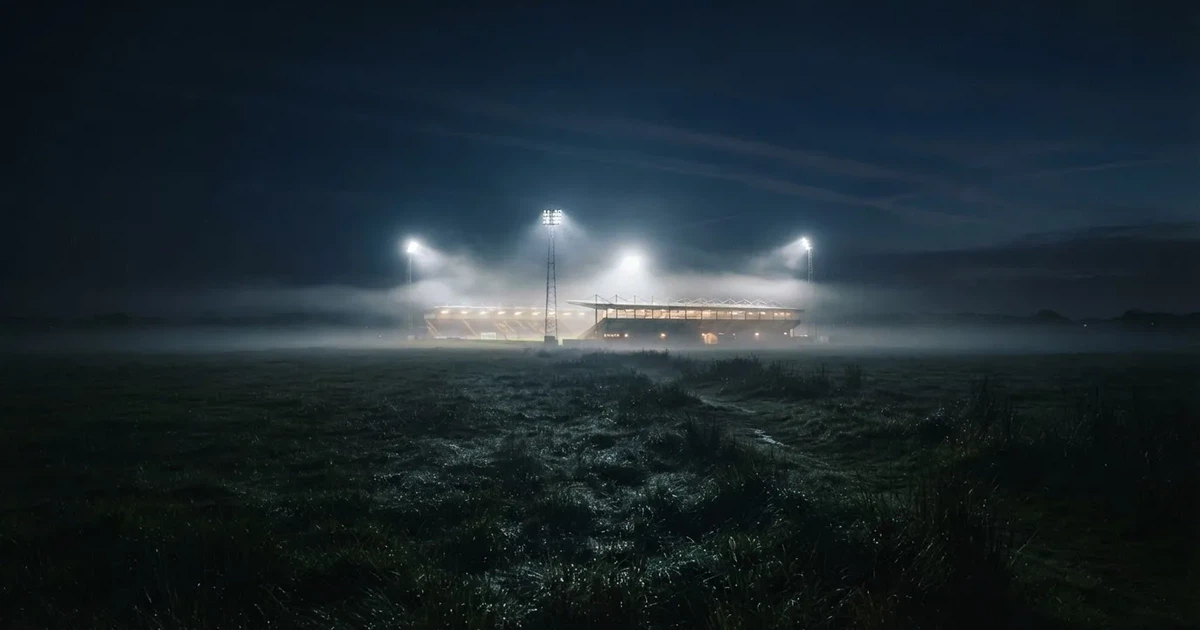 Ein Stadion unter Flutlicht, aus großer Entfernung über eine dunkle Wiese fotografiert, am Himmel zieht Nebel auf.