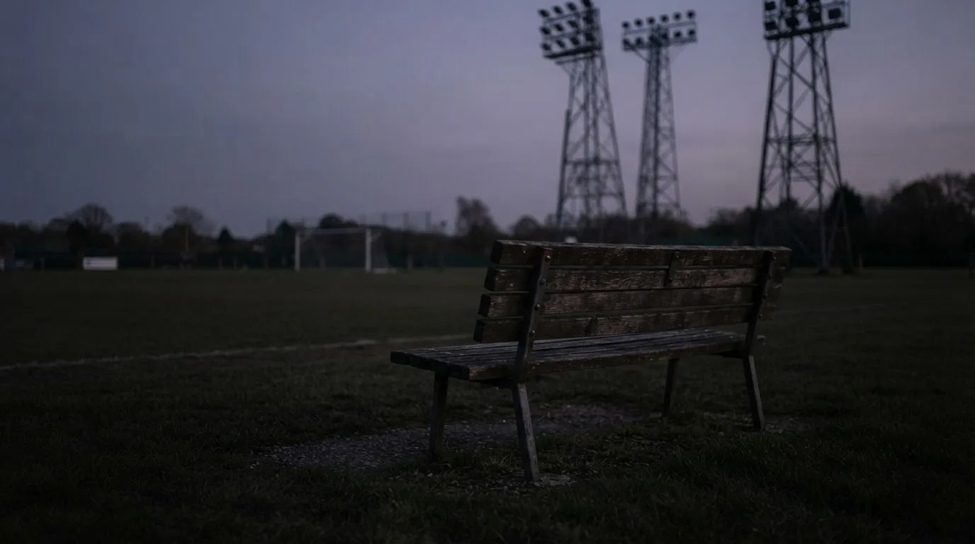 Eine leere Parkbank am Rand eines Fußballfelds in der Abenddämmerung, im Hintergrund ragen dunkle Flutlichtmasten in den Himmel.