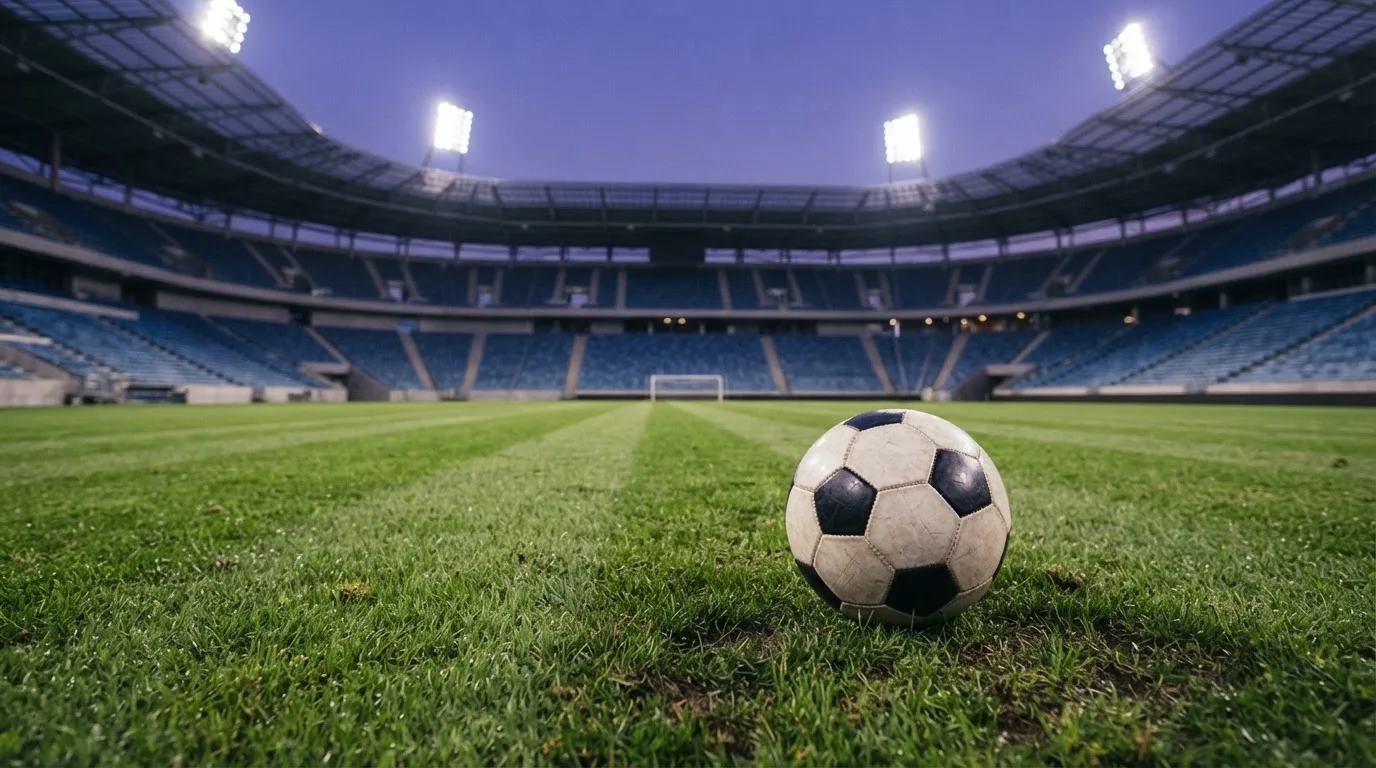 Ein Fußball liegt auf dem Rasen eines leeren Platzes, im Hintergrund ein Stadion unter Flutlicht in der Abenddämmerung.