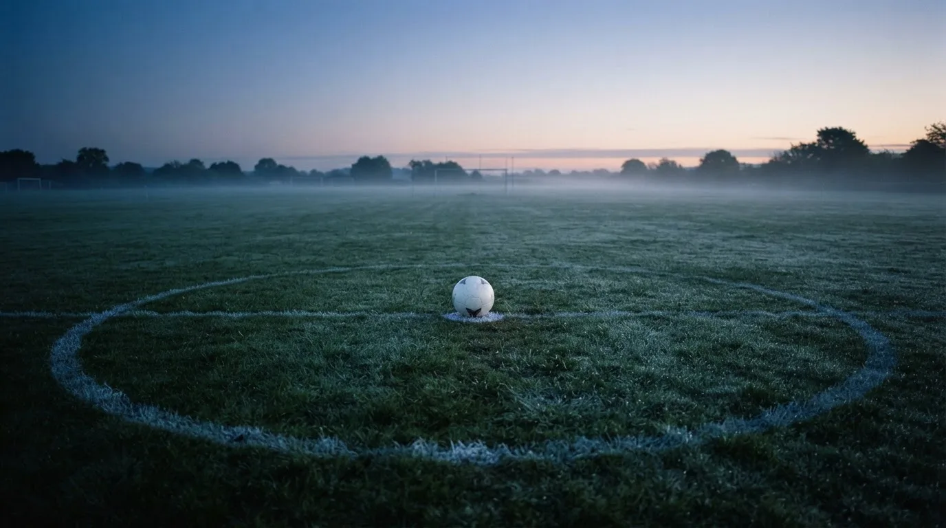 Ein Fußball ruht auf dem Mittelkreis eines leeren Rasenplatzes in der Morgendämmerung, Tau liegt auf dem Gras.