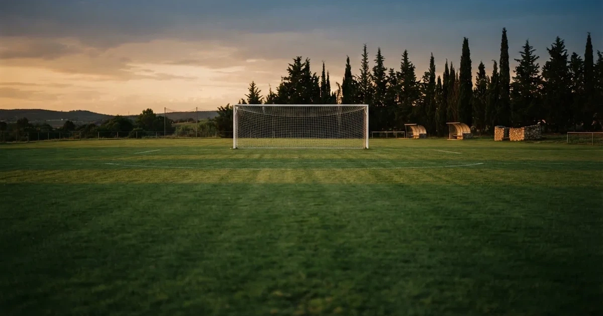 Ein Fußballtor mit Netz auf einem leeren Platz im Abendlicht, der Rasen frisch gemäht, im Hintergrund Zypressen.