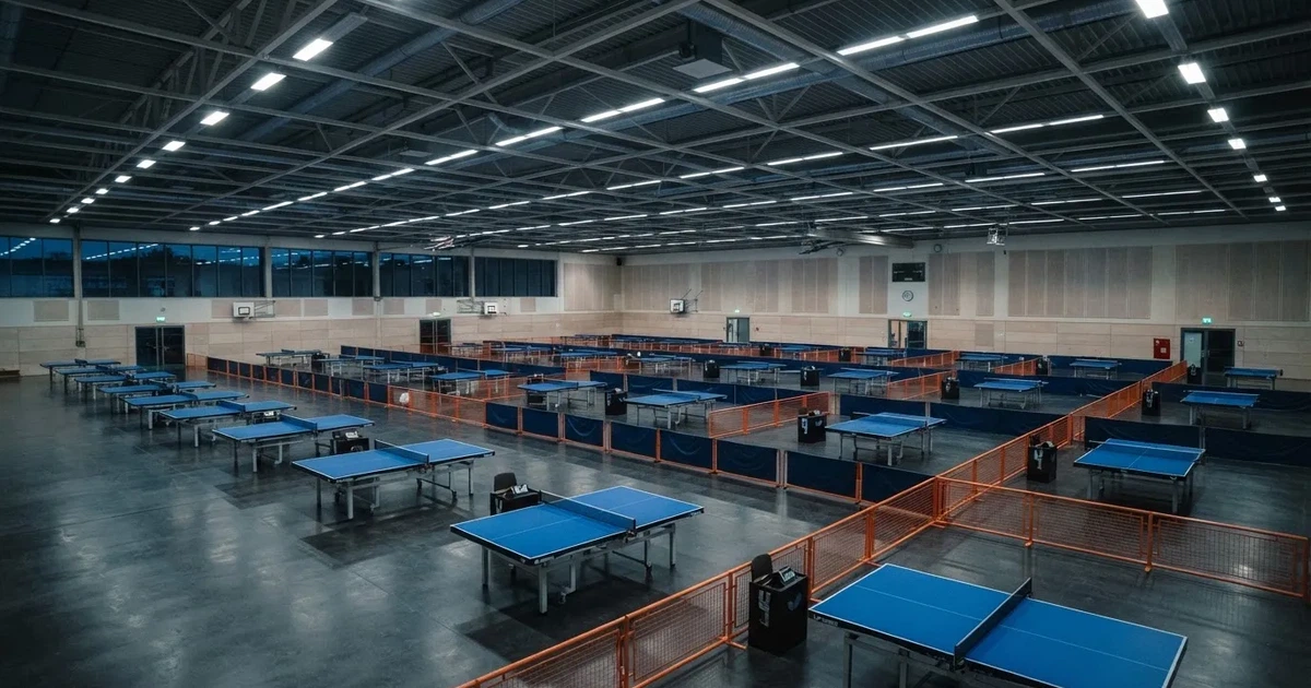 Eine Tischtennishalle aus erhöhter Perspektive mit mehreren blauen Tischen in Reihen unter gedämpftem Hallenlicht, ohne Personen.