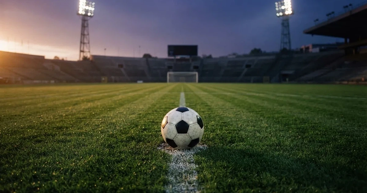 Ein Fußball liegt auf dem Anstoßpunkt eines leeren Rasenplatzes, im Hintergrund eine Tribüne und Flutlichtmasten in der Abenddämmerung.