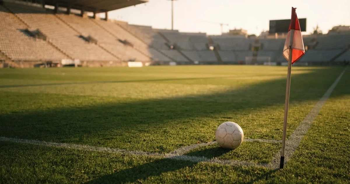 Ein Fußball liegt am Viertelkreis an der Eckfahne eines Rasenplatzes, im Hintergrund leere Tribünen in warmem Abendlicht.