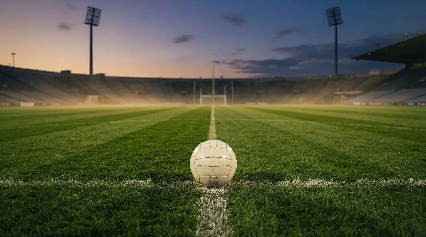 Ein Fußball liegt auf dem Anstoßpunkt eines Rasenplatzes in der Abenddämmerung, Flutlichtmasten stehen unscharf im Hintergrund.