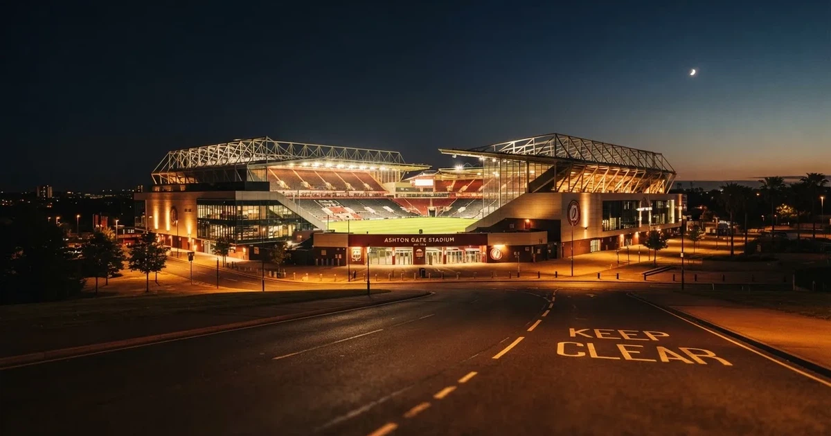 Ein hell erleuchtetes Fußballstadion bei Nacht aus der Ferne gesehen, über dem Dach ein klarer Himmel, davor eine leere Straße.
