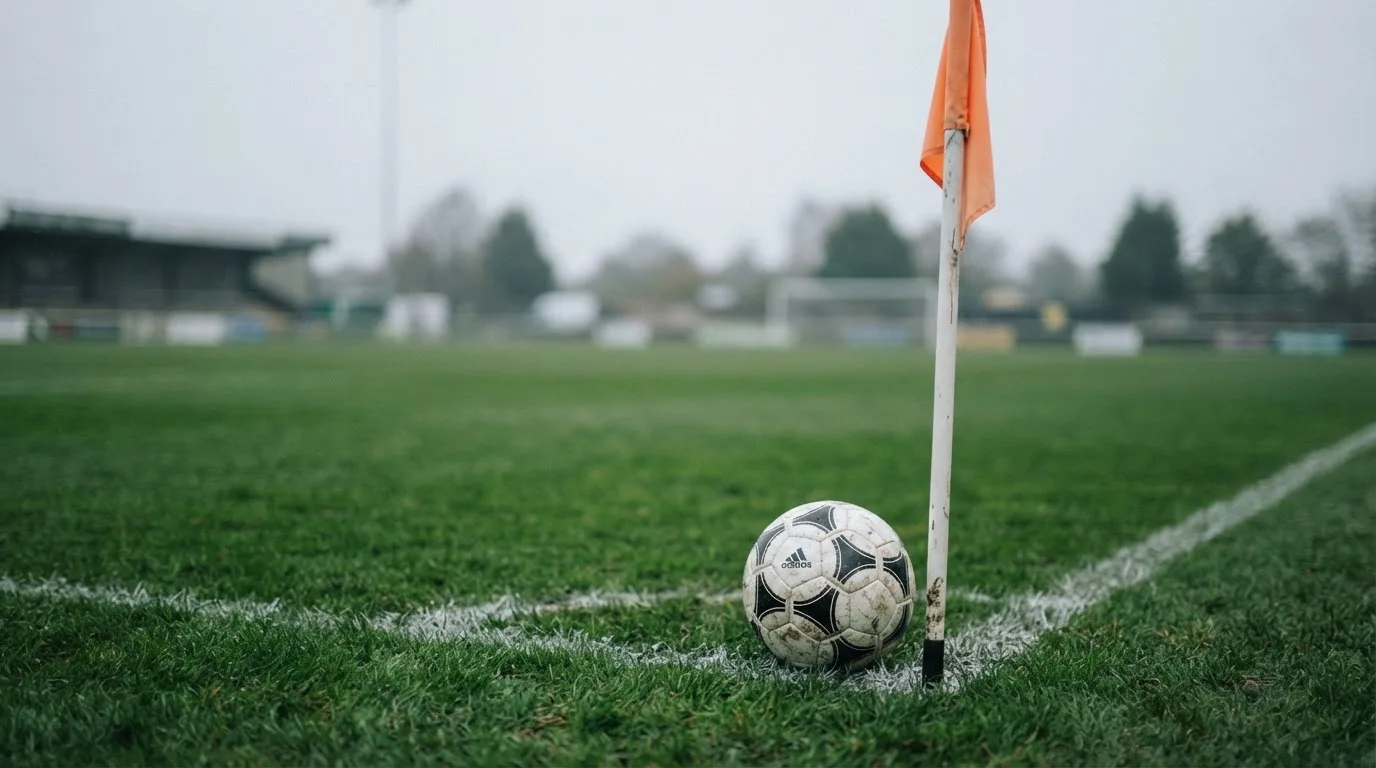 Ein Fußball liegt neben einer Eckfahne auf kurz gemähtem Rasen unter bewölktem Himmel.